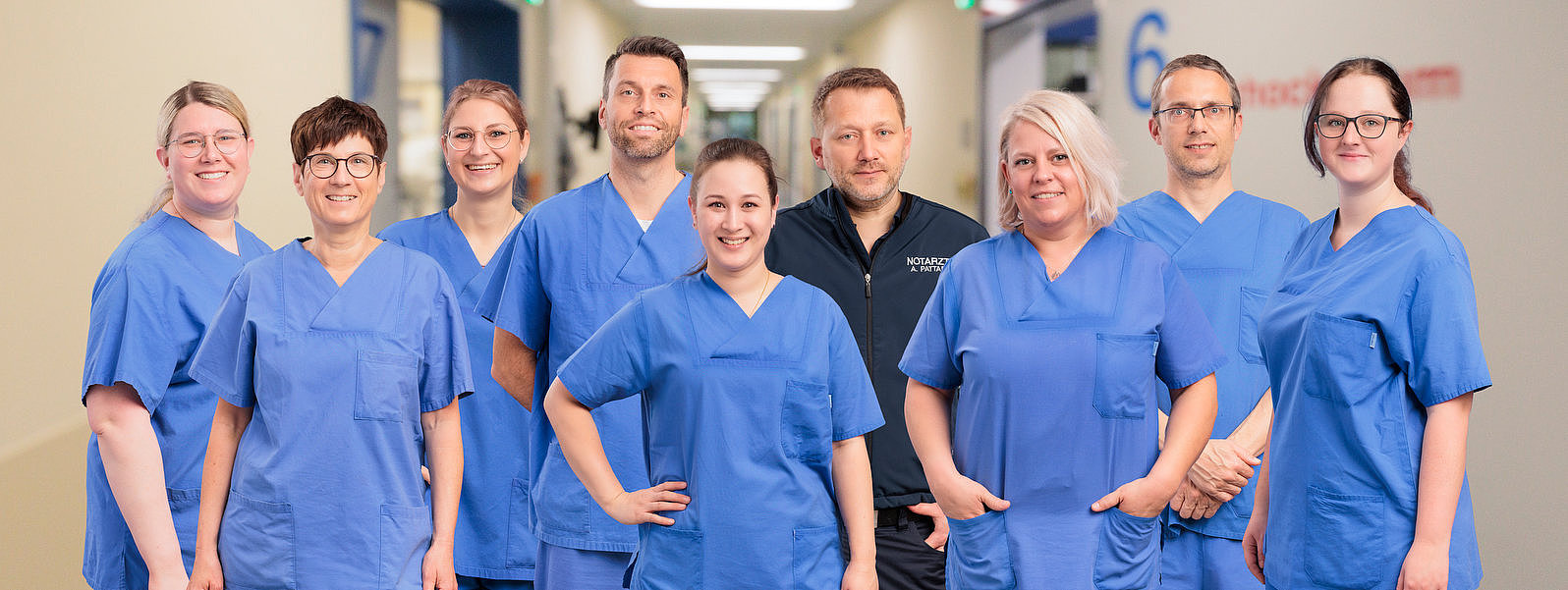 Neun medizinische Fachkräfte in blauer Arbeitskleidung stehen in einem Krankenhausflur und lächeln in die Kamera